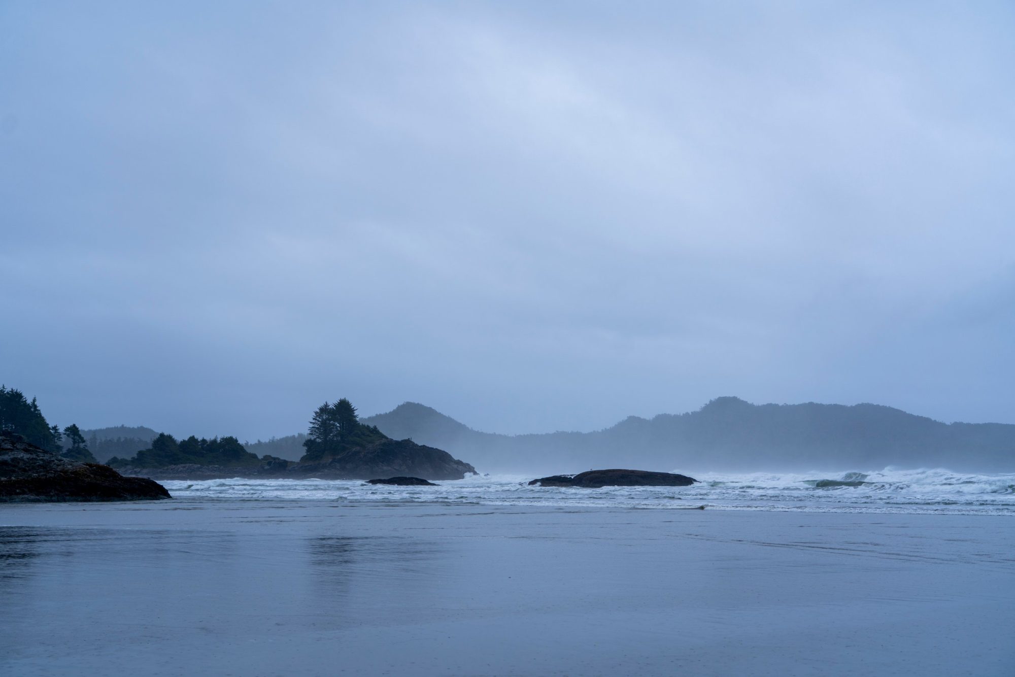 Waves breaking beyond dark rocks on a misty Pacific coast beach